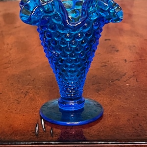 May include: A small, blue glass vase with a ruffled top and a textured, bubbled surface. The vase is on a wooden surface.