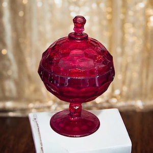 May include: A ruby red glass candy dish with a domed lid and a pedestal base. The dish has a textured, faceted design.