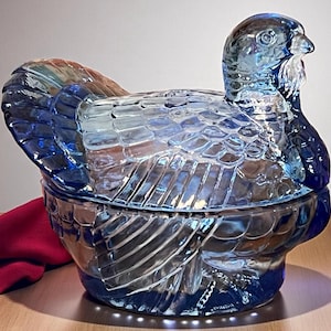 May include: A blue glass turkey-shaped covered dish. The dish has a detailed design with a textured surface, including feathers and a head with a beak. The glass has a reflective quality, and a red cloth is visible in the background.