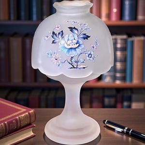 May include: A frosted glass table lamp with a floral design in shades of blue and white. The lamp has a scalloped shade and a pedestal base. A pen and books are on the table. The lamp is approximately 10 inches tall.