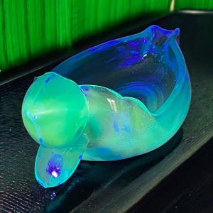 May include: A light blue glass ashtray shaped like a duck. The ashtray has a green iridescent sheen and a blue iridescent rim.