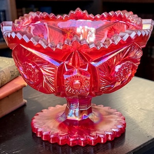May include: A vibrant red carnival glass bowl featuring an iridescent finish and a pedestal base. The bowl has a scalloped rim and is decorated with a pressed pattern of starbursts and geometric shapes across its exterior surface.