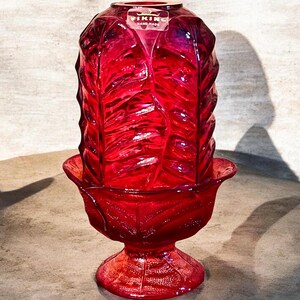 May include: A ruby red Viking hand-made glass candle holder. The candle holder has a leaf-like design and a pedestal base. The top portion is a tall, cylindrical shape, and the base is a flared bowl shape. The Viking logo is visible.
