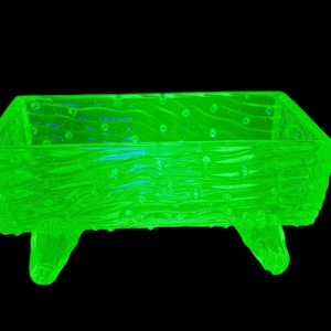 May include: A glowing green rectangular glass dish with a textured, wood-like appearance. The dish is supported by four small, rounded feet and has a dark background.