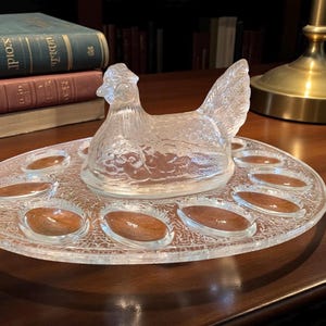 May include: A clear glass hen-shaped egg holder with a matching oval base. The base has spaces for eggs, with a textured design. The hen has detailed feathering and a decorative appearance. The egg holder is on a wooden surface.