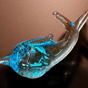 May include: A clear glass dolphin figurine with blue accents. The dolphin has a curved body, a pointed snout, and a split tail. The figurine is approximately 8 inches long and is a decorative item.