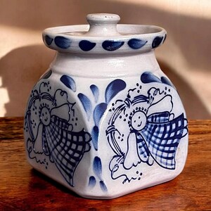 May include: A white ceramic jar with a lid, decorated with blue and white angels and floral designs. The jar has a hexagonal shape and a glossy finish.