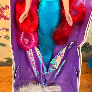 May include: A mermaid doll with long vibrant red hair, wearing a metallic blue bodice and a shimmering purple iridescent tail fin. The doll is packaged in a purple box and includes a small white plastic hairbrush accessory at the bottom.