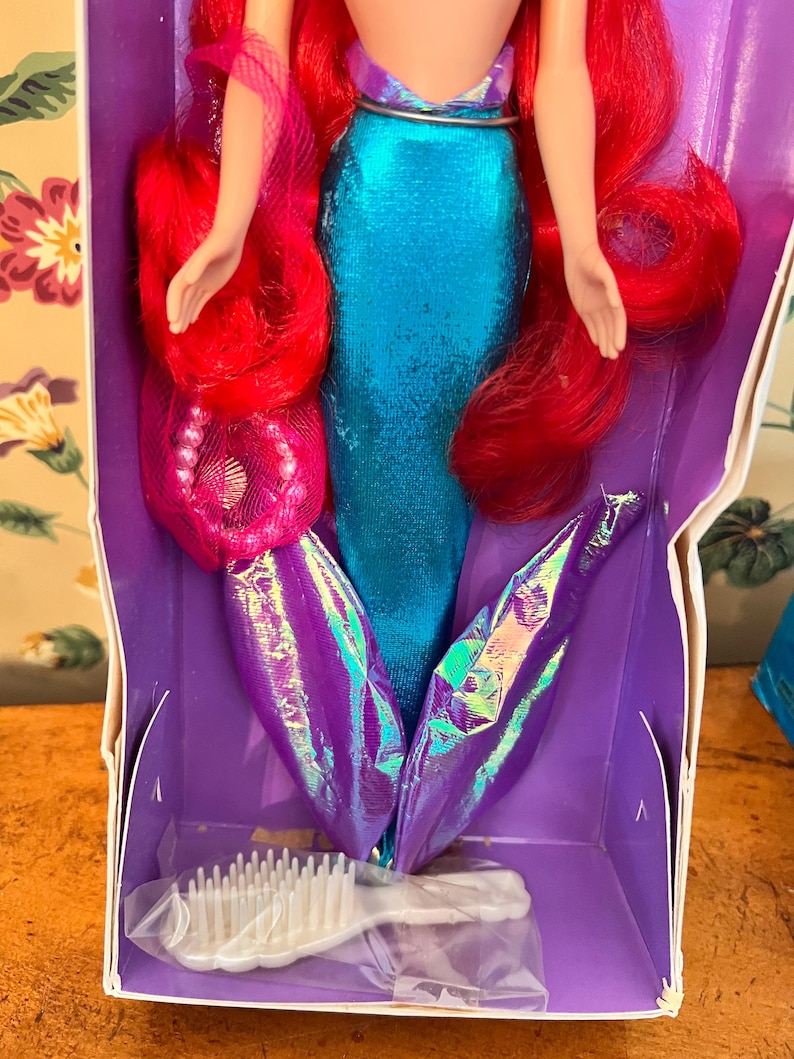 May include: A mermaid doll with long vibrant red hair, wearing a metallic blue bodice and a shimmering purple iridescent tail fin. The doll is packaged in a purple box and includes a small white plastic hairbrush accessory at the bottom.
