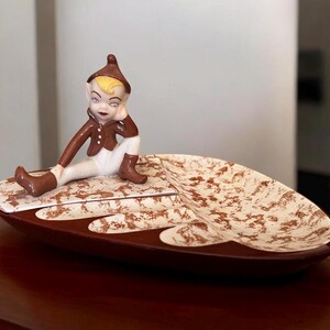 May include: A ceramic leaf-shaped dish with a brown and white speckled glaze. A small figurine of a brown-haired elf sits on the dish, with one hand on their chin.