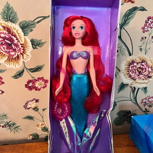 May include: A mermaid doll with long vibrant red hair, a purple seashell-shaped top, and a shimmering teal tail with iridescent purple fins. The doll is packaged in a purple box and includes a small white plastic hairbrush at the bottom.