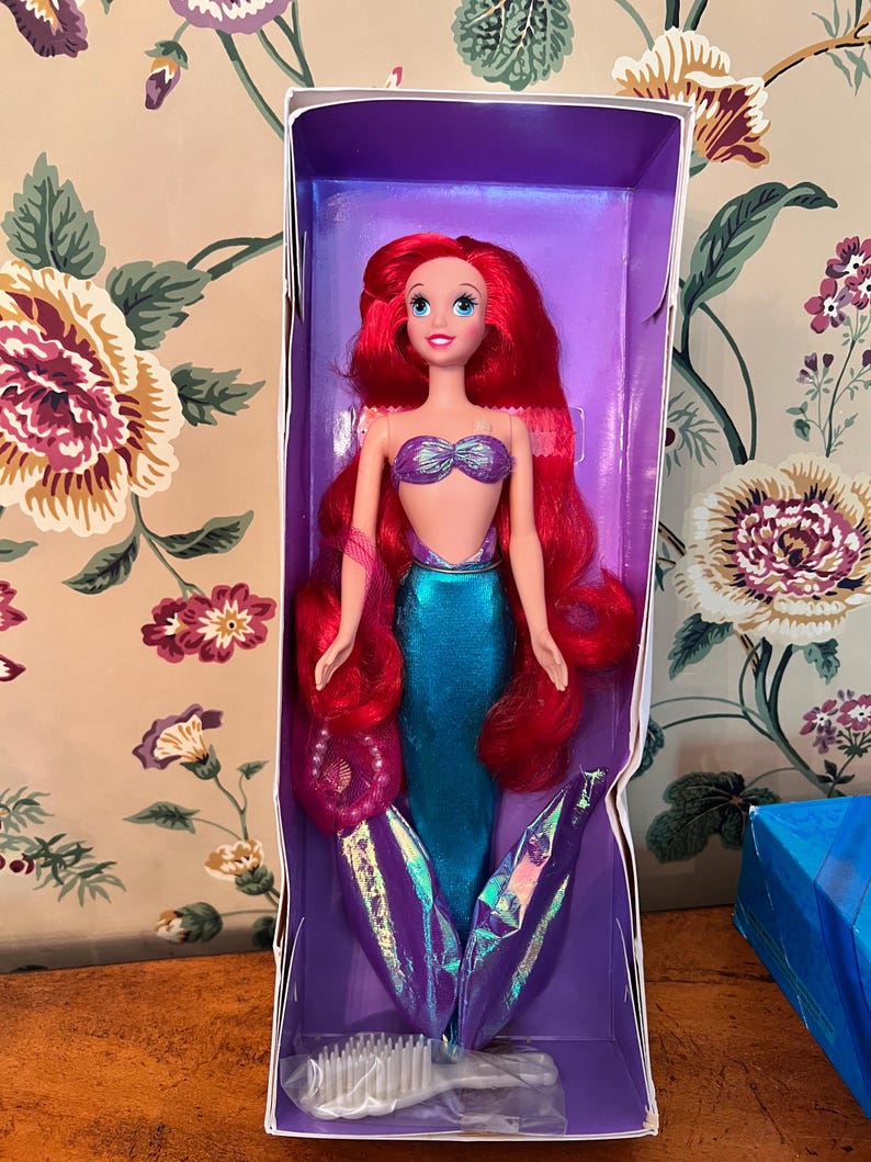 May include: A mermaid doll with long vibrant red hair, a purple seashell-shaped top, and a shimmering teal tail with iridescent purple fins. The doll is packaged in a purple box and includes a small white plastic hairbrush at the bottom.