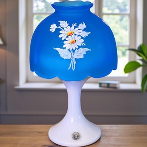 May include: A vintage table lamp with a blue glass shade featuring a white daisy floral design. The lamp has a white base and stem. The shade has a scalloped edge. The lamp is on a wooden surface.