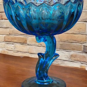 May include: A vibrant blue glass compote with a scalloped rim and a decorative stem. The bowl is supported by a curved, stylized stem and a circular base. The glass has a glossy finish, reflecting light and adding to its visual appeal.