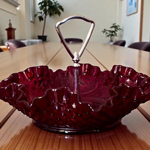 May include: A ruby red, two-tiered serving dish with a ruffled edge and a silver handle. The dish is made of glass and sits on a wooden table. The handle is arched and connects to the center of the dish.