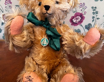 May include: A light brown teddy bear with a green ribbon around its neck. The bear has a stitched nose and pink paw pads. The bear is sitting on a wooden surface and has a tag on its chest that says "Steiff" and "Germany". The bear's foot has the text "Steiff, 1910, Nr 463" on it.