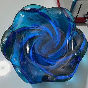 May include: A decorative glass bowl with a swirling blue and teal design. The bowl has a unique, organic shape with a rippled edge. The glass is transparent, allowing light to pass through and highlight the intricate patterns.