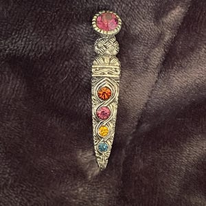 May include: A silver-toned brooch with a decorative design. The brooch features a large pink gemstone at the top and a series of smaller, colorful gemstones in orange, pink, yellow, and blue. The brooch has an ornate, engraved pattern.