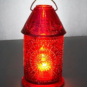 May include: A red glass lantern with a decorative pattern and a metal handle. The lantern is illuminated from within, casting a warm glow. The design features a series of repeating shapes and lines. The lantern is a decorative home accent.