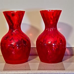 May include: Two red glass vases with a textured, glossy finish. The vases have a bulbous base, a narrow neck, and a flared top. The vases are displayed on a white surface.