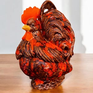 May include: A ceramic rooster figurine in shades of brown and orange. The rooster has a detailed comb, wattle, and tail feathers. The figurine is likely a decorative kitchen item or a collectible piece.