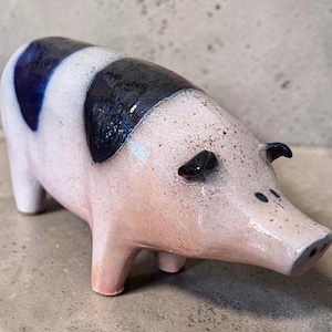 May include: A ceramic pig figurine with a glossy finish, featuring a light pink body with dark blue patches on the back and sides. The snout and face have small dark speckles, and the ears are painted in a solid dark blue color.