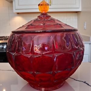 May include: A ruby red, lidded glass jar with a geometric pattern. The jar has a round shape and a lid topped with an orange finial. The jar is sitting on a white surface.