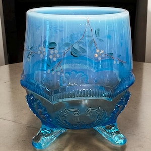 May include: A vintage blue glass bowl with a decorative floral design. The bowl has a flared shape, rests on three ornate feet, and features hand-painted branches with white berries and green leaves.