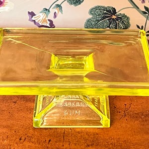 May include: A rectangular, yellow-green glass tray with a raised, square base. The tray has a slightly angled design and the words "TEABERRY GUM" are visible on the base. The tray is likely vintage and sits on a wooden surface.