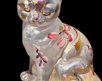 Peut inclure: Figurine de chat en verre irisé avec des motifs floraux roses et violets peints à la main. Le chat assis a les yeux fermés et une expression sereine. La figurine a une finition nacrée, reflétant un arc-en-ciel de couleurs.