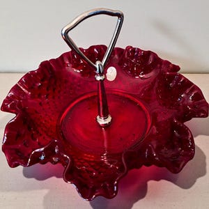 May include: A ruby red glass serving dish with a ruffled edge and a silver metal handle. The dish has a textured surface and a circular base. A small, round sticker is visible on the dish.