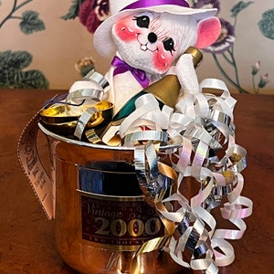May include: A white mouse doll wearing a white top hat with a purple band, holding a bottle of champagne in a copper ice bucket with a gold ribbon and the text "Vintage 2000 Two Thousand".