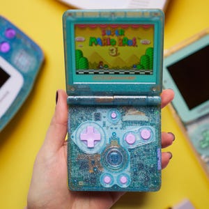 Op de afbeelding: Een blauwgroene glitterende draagbare spelcomputer met een opklapbaar scherm dat het titelscherm van Super Mario Bros. 3 weergeeft. Het apparaat heeft een lichtpaarse richtingsknop, A- en B-knoppen en start- en select-knoppen op de onderkant.