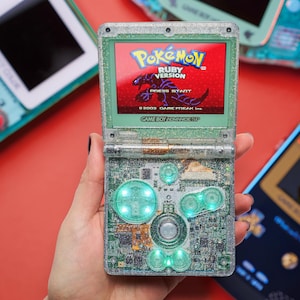 Puede incluir: Una consola portátil Game Boy Advance SP transparente con purpurina y pantalla retroiluminada que muestra la pantalla de título de Pokémon Ruby Version. El dispositivo tiene botones verdes translúcidos y un circuito visible.