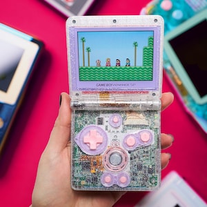 Może przedstawiać: Przezroczysta konsola przenośna Game Boy Advance SP z brokatem i widoczną płytką drukowaną wewnątrz. Urządzenie posiada jasnoróżowe przyciski i krzyżak, a na ekranie wyświetlana jest pikselowa scena z gry wideo z postaciami.