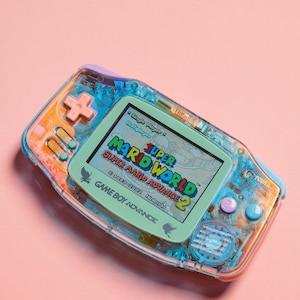May include: A translucent blue and orange Game Boy Advance handheld gaming console with a green screen displaying the game "Super Mario World: Super Mario Advance 2". The game's title is visible on the screen, along with the Nintendo logo and copyright information.