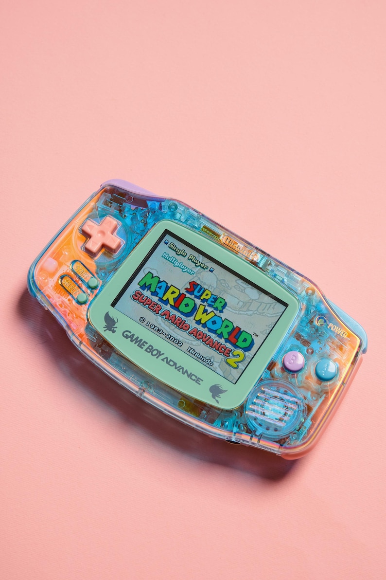May include: A translucent blue and orange Game Boy Advance handheld gaming console with a green screen displaying the game "Super Mario World: Super Mario Advance 2". The game's title is visible on the screen, along with the Nintendo logo and copyright information.