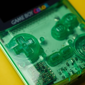 Op de afbeelding: Een doorschijnende limoengroene Game Boy Color handheld spelcomputer met een zichtbare interne printplaat, een richtingspad, A- en B-knoppen, en Select- en Start-knoppen. Het schermframe toont het Game Boy Color logo.