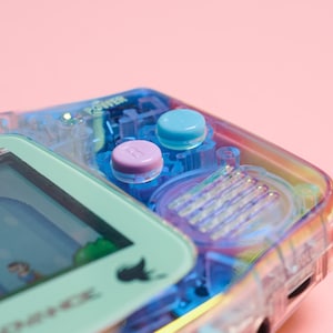 May include: A clear blue and purple handheld video game console with a green screen. The console has two buttons, one blue and one purple.