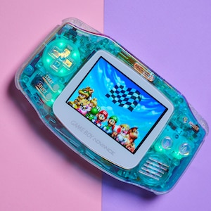 May include: A translucent teal Game Boy Advance handheld gaming console with a white faceplate. The console is displaying a video game featuring Mario and other characters racing on a track. The text "GAME BOY ADVANCE" is printed on the faceplate.