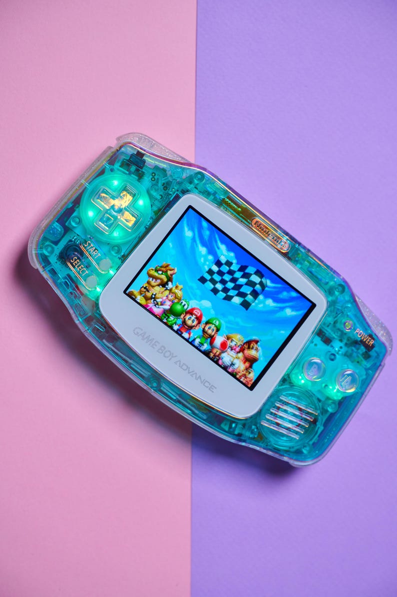 May include: A translucent teal Game Boy Advance handheld gaming console with a white faceplate. The console is displaying a video game featuring Mario and other characters racing on a track. The text "GAME BOY ADVANCE" is printed on the faceplate.