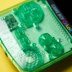 Op de afbeelding: Een draagbare Game Boy Color spelcomputer met een doorschijnende neon-groene plastic behuizing. Het apparaat bevat een richtingspad, A- en B-knoppen, Select- en Start-knoppen en een luidsprekerrooster, allemaal in bijpassend groen uitgevoerd.