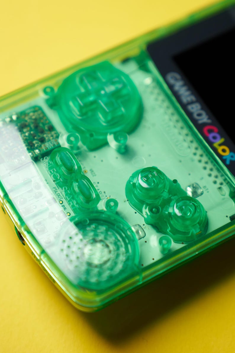Op de afbeelding: Een draagbare Game Boy Color spelcomputer met een doorschijnende neon-groene plastic behuizing. Het apparaat bevat een richtingspad, A- en B-knoppen, Select- en Start-knoppen en een luidsprekerrooster, allemaal in bijpassend groen uitgevoerd.