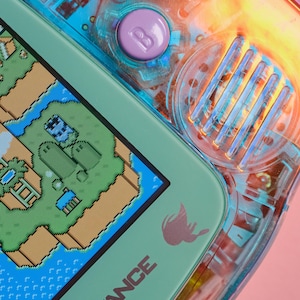 May include: A close-up of a teal Game Boy Advance handheld gaming console with a pixelated game screen showing a green and brown landscape. The console has a translucent blue and orange shell with a purple button labelled "B" and a blue button labelled "A". The text "Game Boy Advance" is printed on the bottom of the console.