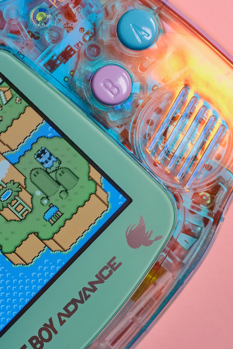 May include: A close-up of a teal Game Boy Advance handheld gaming console with a pixelated game screen showing a green and brown landscape. The console has a translucent blue and orange shell with a purple button labelled "B" and a blue button labelled "A". The text "Game Boy Advance" is printed on the bottom of the console.