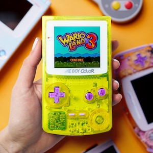 Pode incluir: Uma consola portátil Game Boy Color amarela néon transparente exibindo o jogo "Wario Land 3". A consola tem botões roxos e um D-pad. Outras consolas de jogos retro estão no fundo.