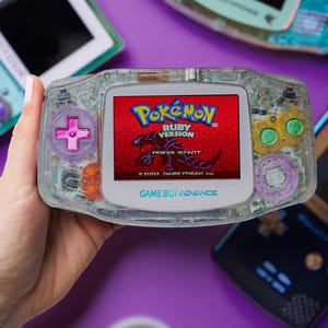 May include: A clear Game Boy Advance handheld console with a Pokemon Ruby Version game displayed on the screen. The console has translucent purple buttons and a clear shell. Other consoles and a controller are visible on a purple surface.
