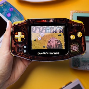 Könnte beinhalten: Ein Game Boy Advance Handheld-Konsolen mit einer transparenten braunen Hülle und gelben Akzenten. Der Bildschirm zeigt eine farbenfrohe Videospielszene. Weitere Retro-Spielkonsolen sind im Hintergrund zu sehen.