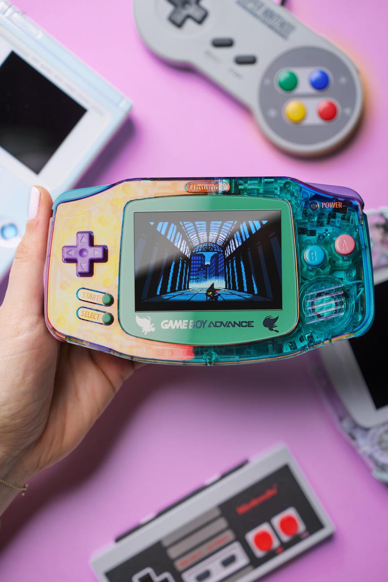 May include: A teal, green and yellow Game Boy Advance handheld gaming console with a colourful, abstract design. The screen shows a video game with a dark figure walking through a futuristic city. The text "GAME BOY ADVANCE" is printed on the console.