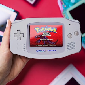 May include: A grey Game Boy Advance console showing the "Pokémon Ruby Version" game on its screen. The console has a directional pad and buttons. The text "GAME BOY ADVANCE" is visible. Other retro consoles are in the background, all on a red surface.